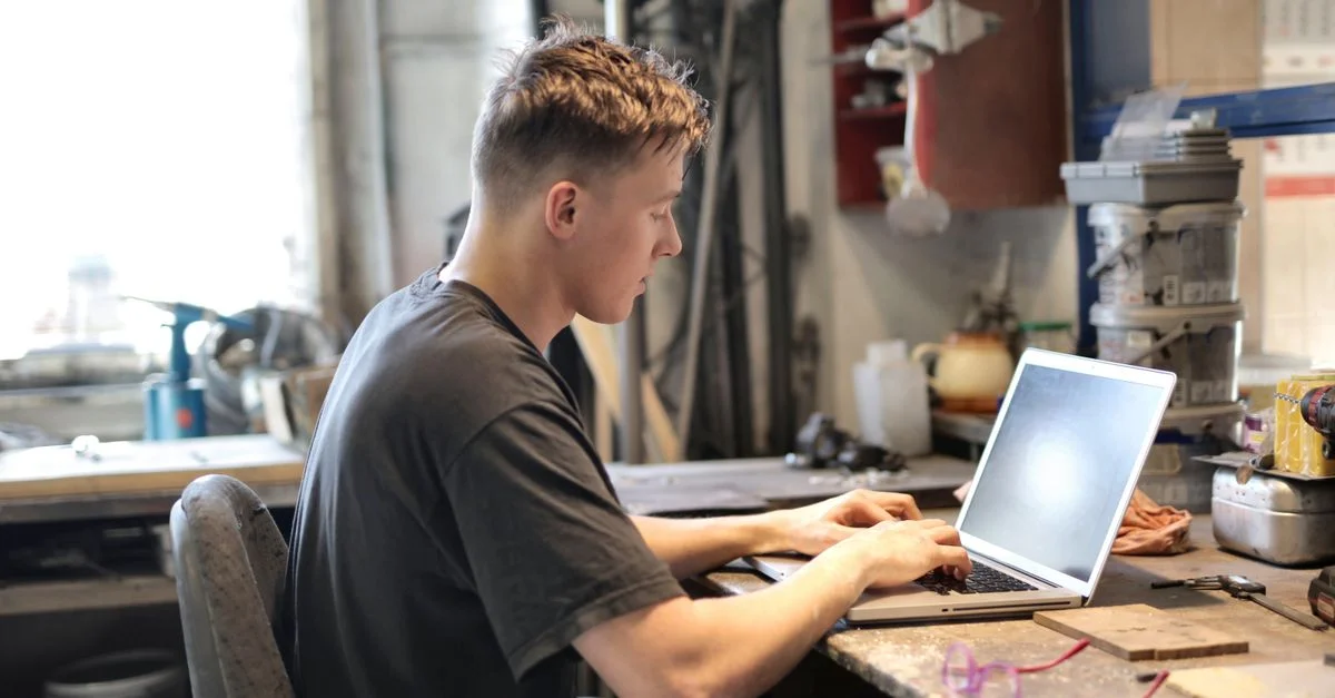 Homme concentré travaillant à distance depuis un atelier de garage avec un ordinateur portable.