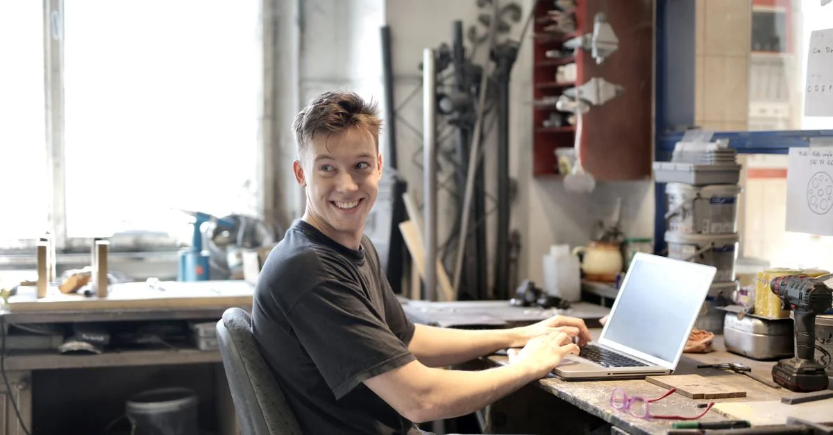 Jeune homme souriant utilisant un ordinateur portable dans un espace de travail créatif avec des outils autour.